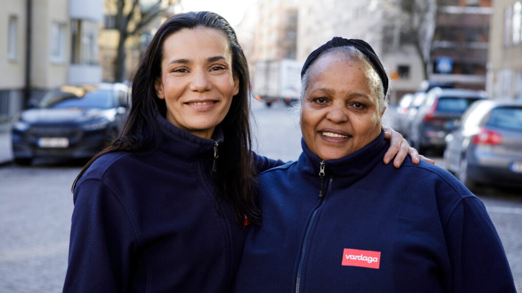 Två kvinnliga medarbetare i arbetskläder från Vardaga står tillsammans utomhus och ler mot kameran.