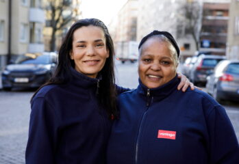 Två kvinnliga medarbetare i arbetskläder från Vardaga står tillsammans utomhus och ler mot kameran.